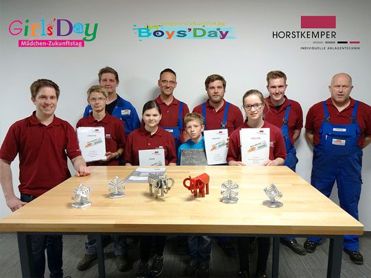 A group of teenagers and adults in work clothes standing behind a table with technical models and certificates at Horstkemper Maschinenbau during the student internship for Girls' Day and Boys' Day 