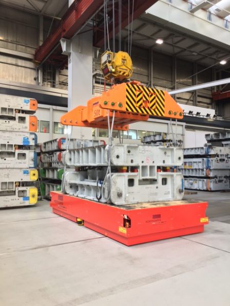 Industrielle Hebevorrichtung mit gelb-orangefarbenem Magnetgreifer hebt schwere graue Metallmaschine in einer Werkhalle mit Regalen im Hintergrund