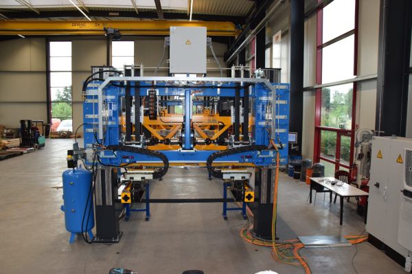 Industrielle Transfersystemanlage mit blauer Rahmenkonstruktion, gelben Greifern und Steuerungseinheiten in einer Werkhalle mit großen Fenstern