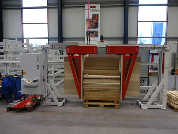 Ausrichtstation mit rotem Hebezeug über einer Palette mit Holzplatten in einer Halle mit großen Fenstern