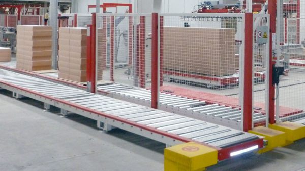 Industrial production line featuring a conveyor belt, automated gripper arms, and stacked boxes in a large warehouse