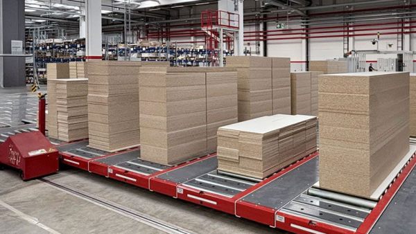 Blick auf ein Fördertechniksystem mit Rollenbahnen und mehreren gestapelten Holzplattenpaketen in einer Produktionshalle.