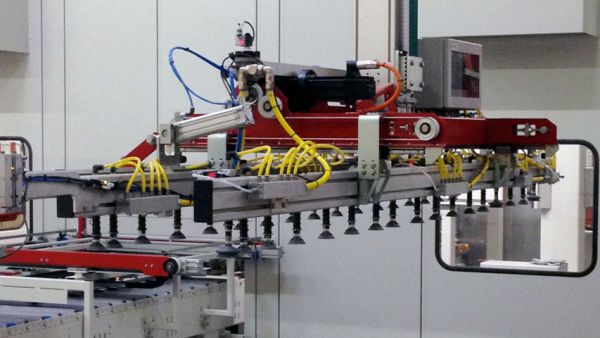 Gantry feeder system with a red gripper arm and yellow hoses above a conveyor belt carrying stacked wooden panels in an industrial warehouse 