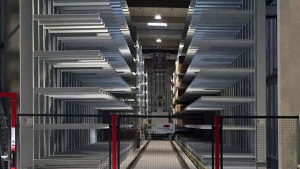 Front view of a high-bay warehouse with storage bays on both sides. The stacker crane is located in the center of the aisle between the rows of shelves.