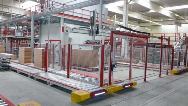 Industrielle Anlagenverkettung mit Förderband, automatisierten Greifarmen und gestapelten Kartons in einer großen Halle