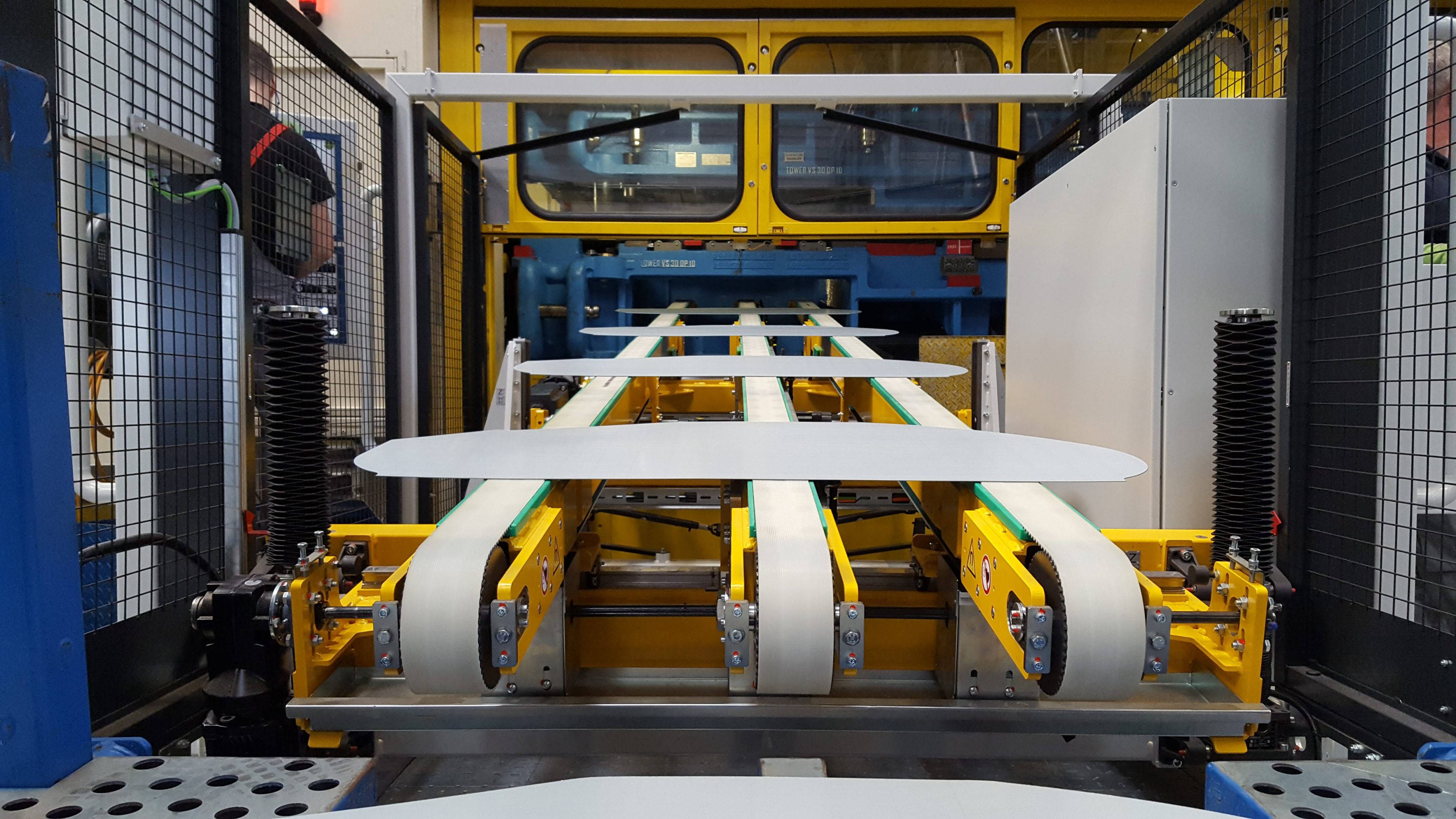 Blick auf eine gelb lackierte Stapelanlage mit mehreren parallelen Förderbändern und weißen Platten darauf in einer Industrieumgebung