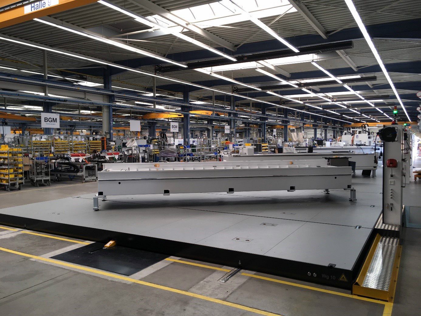 Overall view of a flow and cycle assembly line in a production hall. Several assembly stations are arranged along the conveyor line, while workpiece carriers are on the line.